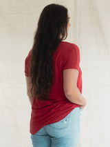 A woman with long dark hair is shown from behind wearing a Poppy red Blondie Janis t-shirt and light blue jeans, standing against a neutral off-white fabric backdrop.