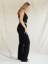 A woman stands in profile wearing a black sleeveless crop top and matching black Blondie Juniper pant against a draped off-white fabric backdrop.