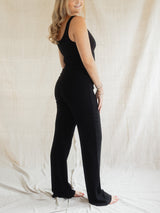 A woman modeling a black sleeveless jumpsuit with Blondie Juniper pant, standing against a draped off-white fabric backdrop.