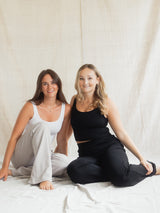 Two women sitting side-by-side on a white sheet against a beige fabric backdrop. The woman on the left wears a white tank top and light gray Blondie Juniper pant, while the woman on the right wears a black tank top and black Blondie Juniper pant.