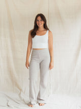 A woman stands barefoot in front of a draped cream fabric backdrop, wearing a white cropped tank top and light heather Flax gray Blondie Juniper pant.