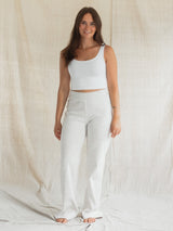 A woman stands barefoot in front of a draped beige fabric backdrop, wearing a white ribbed tank top and white Confetti textured Blondie Juniper pant.