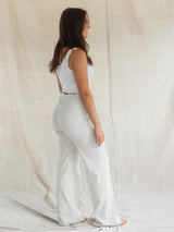 A woman stands in profile wearing a white sleeveless top and textured white Confetti Blondie Juniper pant against a draped cream fabric backdrop.