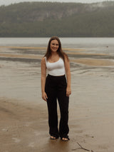 A woman stands on a sandy beach with water and a forested hill in the background, wearing a white tank top and black Blondie Juniper pant.
