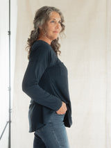A woman with gray curly hair is shown in profile wearing a dark navy blue Blondie Long-Sleeve Janis T-shirt and blue jeans, standing against a light beige fabric backdrop.