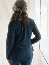 A woman viewed from behind wearing a dark navy blue Blondie Long-Sleeve Janis T-shirt and blue jeans, standing in front of a bright white curtain.
