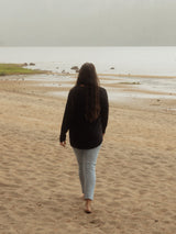 A woman with long dark hair walks barefoot on a sandy beach, viewed from behind, wearing a black Blondie Long-Sleeve Janis T-shirt and light blue jeans, with water and a hazy shoreline in the background.