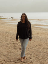 A woman stands barefoot on a sandy beach wearing a black Blondie Long-Sleeve Janis T-shirt and light blue jeans, with the ocean and overcast sky in the background.