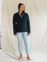 A woman stands barefoot in front of a draped beige fabric backdrop, wearing a dark navy quarter-zip long-sleeve Blondie Mountaineer Sweater with white piping and light-wash blue jeans.
