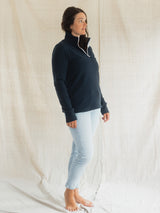 A woman modeling a dark navy quarter-zip Blondie Mountaineer Sweater with a white drawstring, paired with light-wash skinny jeans, standing against a draped off-white fabric backdrop.