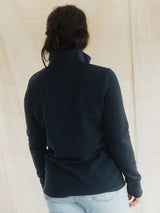 A person shown from the back wearing a dark navy long-sleeve quarter-zip Blondie Mountaineer Sweater and light blue jeans, standing against a neutral beige fabric backdrop.