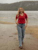 A woman stands on a sandy shoreline with water and forested hills in the background, wearing a fitted Poppy red short-sleeve Blondie New Rideau Shoreline t-shirt and light blue jeans.
