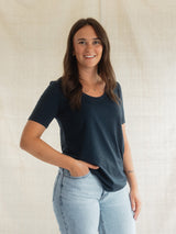 A woman with long brown hair smiling, wearing a dark navy blue short-sleeve Blondie New Rideau Shoreline t-shirt and light wash blue jeans, standing against a neutral beige fabric backdrop.