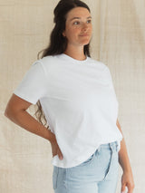A woman wearing a plain white short-sleeve crewneck Blondie Apparel Prairie t-shirt and light blue jeans, standing against a neutral beige textured fabric backdrop.