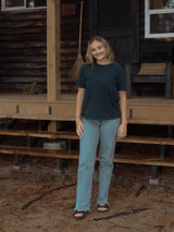A woman stands on the porch of a rustic log cabin, wearing a dark teal short-sleeve Blondie prairie t-shirt, light blue straight-leg jeans, and black sandals.
