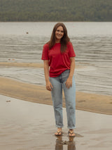 A woman stands on a sandy shoreline with calm water and forested hills in the background, wearing a Blondie Apparel Poppy red short-sleeve Prairie tee shirt and light blue jeans.