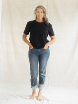A woman with gray hair stands barefoot against a white fabric backdrop, wearing a black short-sleeve Blondie Apparel Prairie t-shirt and light blue cuffed jeans.