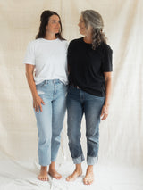 Two women standing barefoot against a draped cream fabric backdrop, wearing short-sleeve Blondie Apparel Prairie t-shirts and jeans. The woman on the left wears a white t-shirt and light-wash blue jeans, while the woman on the right wears a black Blondie Apparel Prairie t-shirt and dark-wash blue jeans.