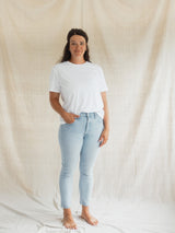 A woman stands barefoot in front of a draped beige fabric backdrop, wearing a plain white short-sleeve Blondie Apparel Prairie T-shirt and light-wash blue skinny jeans.