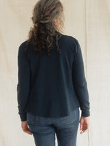A woman viewed from behind wearing a dark navy blue long-sleeve Blondie Spurce Cardigan and blue jeans, standing against a light beige fabric backdrop.