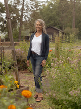 A smiling woman with gray hair stands in a garden path wearing a white t-shirt, dark navy Blondie Spurce Cardigan, blue jeans, and brown sandals. The background features lush green foliage, a wooden structure, and orange flowers in the foreground.