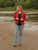 A woman stands on a sandy beach next to a body of water, wearing a Poppy red Blondie Spurce Cardigan, black top, and light blue jeans, with a forest in the background.