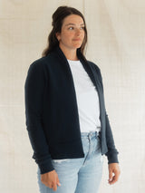 A woman wearing a dark navy blue open-front Blondie Spruce Cardigan over a white t-shirt and light-wash blue jeans, standing against a neutral light beige fabric backdrop.