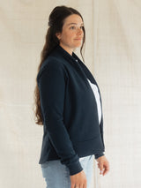 A woman wearing a dark navy blue Blondie Spruce Cardigan over a white top and light blue jeans, standing against a neutral off-white textured backdrop.