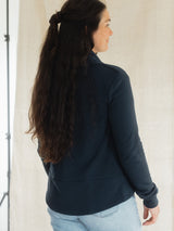 A woman wearing a dark navy blue Blondie Spruce Cardigan over a white top and light blue jeans, standing against a neutral off-white textured backdrop.