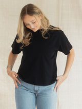 A woman wearing a black short-sleeve Blondie tide tee in slub cotton fabric and light blue jeans, standing against a neutral beige textured backdrop.