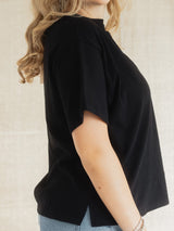 A woman wearing a black short-sleeve Blondie tide tee in slub cotton fabric with a high mock neck and side slits, paired with light blue jeans, shown from a side profile against a neutral beige background.