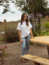 A woman stands outdoors at a wooden picnic table near a garden arch and flowers, wearing a loose white short-sleeve Blondie Tide Tee in a waffle fabric and light blue jeans.