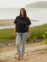 A woman stands barefoot on a sandy shore wearing a loose Navy Blue Blondie Tide Tee in a waffle fabric and light-wash blue jeans, with grass, water, and a distant shoreline in the background.