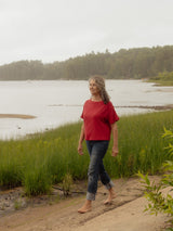 A woman walking barefoot on a sandy path next to a grassy shoreline with a calm lake and forest in the background, wearing a red short-sleeve Blondie Tide Tee in a waffle fabric and dark cuffed jeans.
