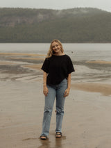 A young woman stands on a sandy shoreline with water and a forested hill in the background, wearing a loose black short-sleeve Blondie Tide Tee in a waffle fabric, light blue jeans, and sandals.