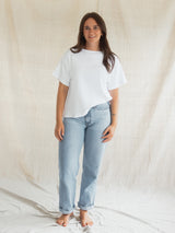 A woman stands barefoot against a draped beige fabric backdrop, wearing a loose white short-sleeve Blondie Tide Tee in a waffle fabric and light blue straight-leg jeans.