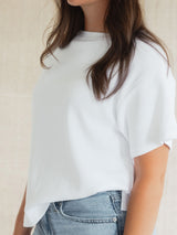 A close-up of a woman wearing a white short-sleeve Blondie Tide Tee in a waffle fabric with a side-slit hem, paired with light-wash blue jeans against a neutral beige background.