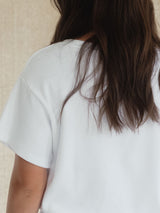A close-up back view of a person with long dark brown hair wearing a white short-sleeve ribbed Blondie Tide Tee in a waffle fabric against a neutral beige textured background.