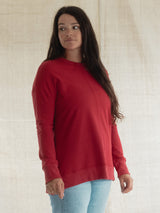A woman wearing a red long-sleeve crewneck Blondie Wheat Sweater and light blue jeans, standing against a neutral beige fabric backdrop.