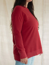 A woman wearing a red long-sleeve crewneck Blondie Wheat Sweater, shown from a side profile against a light beige textured fabric backdrop.