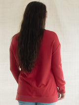 A woman with long, wavy dark brown hair is shown from behind, wearing a red long-sleeve Blondie Wheat Sweater and light blue jeans against a neutral, textured off-white background.