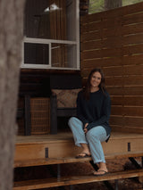 A woman sits on wooden steps in front of a rustic cabin, wearing a dark navy long-sleeve Blondie Wheat Sweater and light blue jeans.