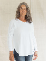A woman with gray hair wearing a white long-sleeve Blondie Willow Sweater and blue jeans, standing against a light beige textured fabric backdrop.