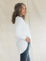 A woman with curly gray-brown hair is shown in profile wearing a loose white long-sleeve Blondie Willow Sweater and dark blue jeans, standing against a light beige fabric backdrop.