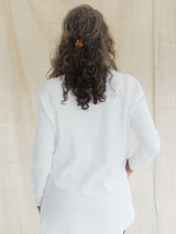 A person viewed from behind wearing a plain white long-sleeve Blondie Willow Sweater, standing against a light beige fabric backdrop. The hair is dark brown and curly, held back with a small orange hair clip.