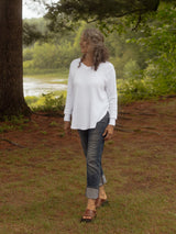 A woman wearing a white long-sleeve Blondie Willow Sweater, cuffed blue jeans, and brown sandals walking outdoors on a grassy area near a tree and water.