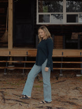 A woman wearing a dark teal long-sleeve Blondie Willow Sweater and light blue flare jeans walks in front of a rustic wooden cabin porch.