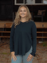 A smiling blonde woman wearing a dark teal long-sleeve knit Blondie Willow Sweater and light blue jeans, standing in front of a rustic wooden structure.