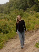 A woman walking on a dirt path outdoors, wearing a black long-sleeve Blondie Willow Sweater and light blue jeans, surrounded by green bushes and trees.