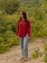 A woman wearing a red long-sleeve Blondie Willow Sweater and light blue jeans stands on a dirt path surrounded by green bushes and foliage.